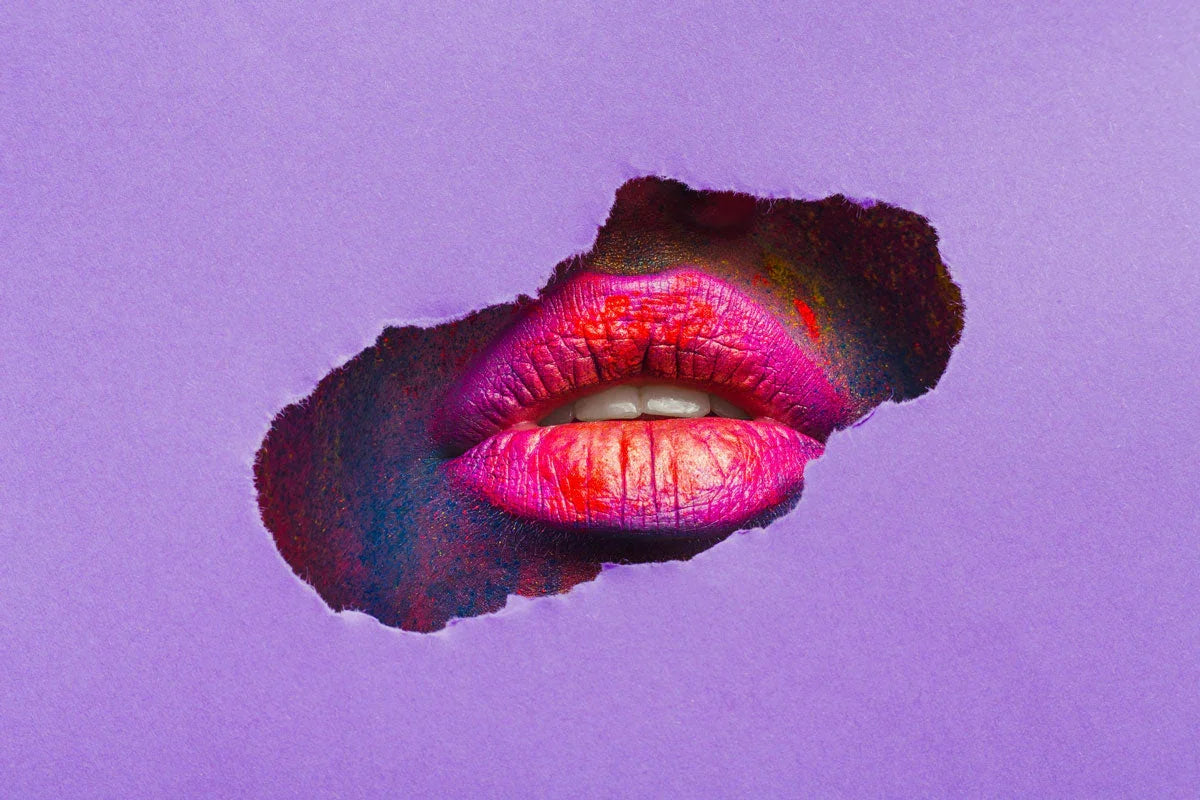 Close-up of vibrant pink and red glittery lips appearing through a tear in purple paper.